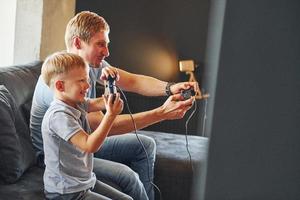 Holding video game controllers. Father and son is indoors at home together photo