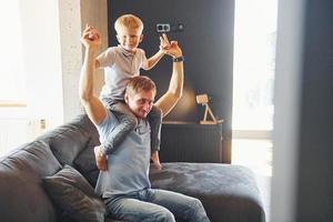 niño está sentado sobre los hombros del hombre. padre e hijo están juntos en casa en el interior foto