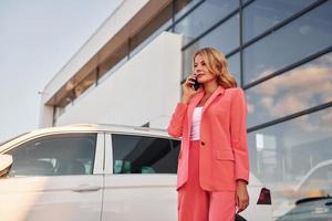 Having conversation by phone. Woman in formal wear standing outdoors with phone in hands and near car photo