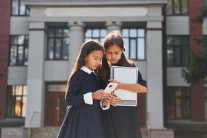 vista frontal. dos colegialas están afuera juntas cerca del edificio de la escuela foto