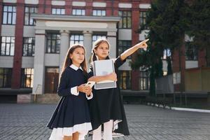 Holding books. Two schoolgirls is outside together near school building photo