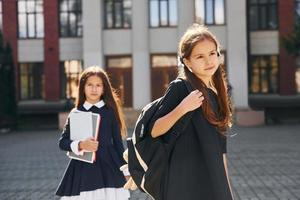 vista frontal. dos colegialas están afuera juntas cerca del edificio de la escuela foto