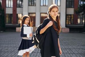 vista frontal. dos colegialas están afuera juntas cerca del edificio de la escuela foto