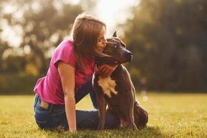 On the green grass. Woman in casual clothes is with pit bull outdoors photo