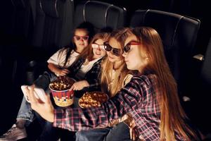 Holding phone in hand. Group of kids sitting in cinema and watching movie together photo