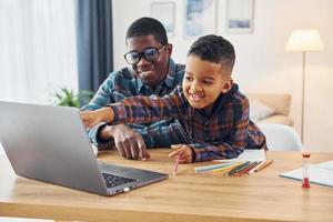 con laptop en la mesa. padre afroamericano con su hijo pequeño en casa foto