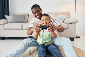 usando joysticks para jugar videojuegos. padre afroamericano con su hijo pequeño en casa foto