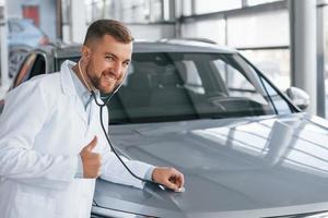 Showing thumb up. Medic in white coat is using stethoscope on the automobile photo