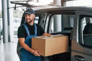 With box in hands. Delivery man in uniform is indoors with car and with order photo