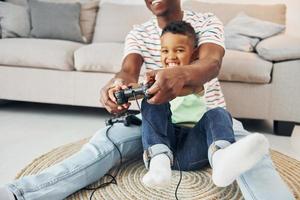 con joysticks en las manos. padre afroamericano con su hijo pequeño en casa foto