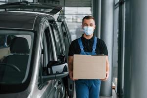 In mask. Delivery man in uniform is indoors with car and with order photo