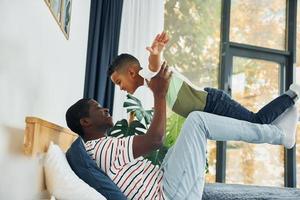 Laying down on the bed and having fun. African american father with his young son at home photo