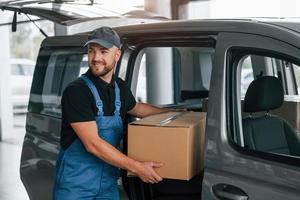 With box in hands. Delivery man in uniform is indoors with car and with order photo