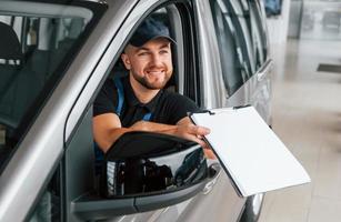 Sitting in vehicle and holding document. Delivery man in uniform is indoors with car and with order photo