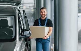 In blue colored clothes. Delivery man in uniform is indoors with car and with order photo