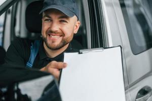 Cheerful worker. Delivery man in uniform is indoors with car and with order photo