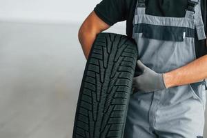Man in uniform is working in the autosalon at daytime. Holding tire in hands photo