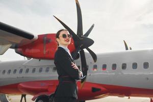Young stewardess that is in formal black clothes is standing outdoors near plane photo