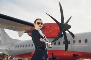 una joven azafata que está vestida de negro formal está parada al aire libre cerca del avión foto