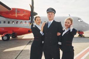 piloto y dos azafatas. tripulación de trabajadores del aeropuerto y del avión con ropa formal de pie juntos al aire libre foto