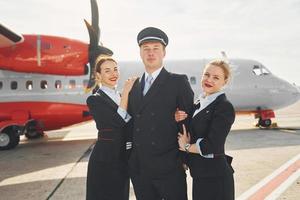 piloto y dos azafatas. tripulación de trabajadores del aeropuerto y del avión con ropa formal de pie juntos al aire libre foto