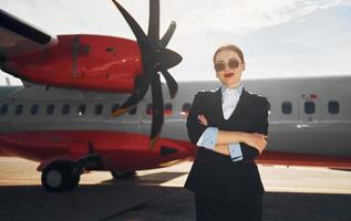 una joven azafata que está vestida de negro formal está parada al aire libre cerca del avión foto