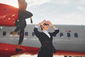 mirando lejos una joven azafata que está vestida de negro formal está parada al aire libre cerca del avión foto