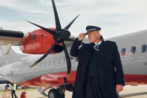 Looking far away. Pilot in formal black uniform is standing outdoors near plane photo