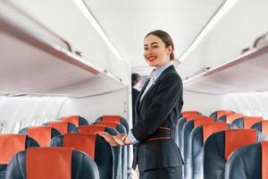 Young stewardess that is in formal black clothes is standing indoors in the plane photo