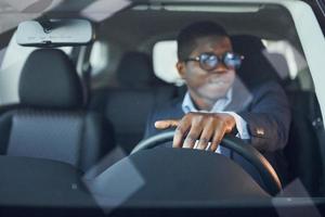 Front view. Young african american businessman in black suit is in the automobile photo