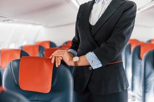 Young stewardess that is in formal black clothes is standing indoors in the plane photo