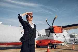 At daytime. Young stewardess that is in formal black clothes is standing outdoors near plane photo