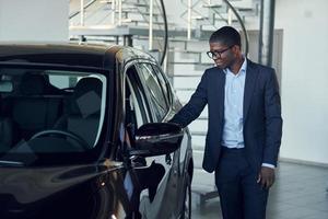 Looking at car. Young african american businessman in black suit is the autosalon photo