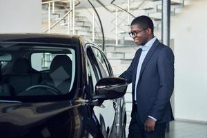 de pie cerca del coche. joven empresario afroamericano en traje negro es el autosalon foto