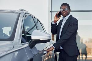 un joven hombre de negocios afroamericano con traje negro está cerca de un automóvil al aire libre foto