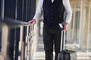 vista de cerca del pasajero masculino con ropa elegante y formal que está en la sala del aeropuerto con equipaje y boletos foto