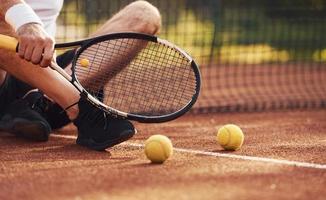 Sitting and taking a break. Close up view of senior man in white shirt and black sportive shorts that is on tennis court photo