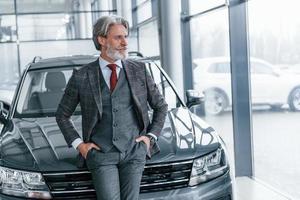 Senior businessman in suit and tie with gray hair and beard standing indoors near car photo