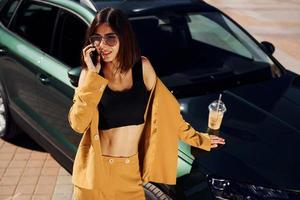 With smartphone. Young fashionable woman in burgundy colored coat at daytime with her car photo