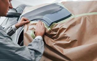 Covering car by brown cloth. Caucasian automobile repairman in uniform works in garage photo