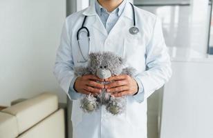 With teddy bear. Professional medic in white coat is in the clinic photo