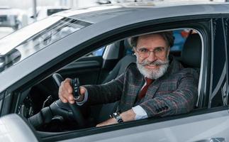 Modern stylish senior man with grey hair and mustache holding keys in modern car photo