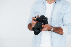 Holds camera in hands. Close up particle view of young man that standing indoors against white background photo