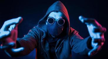Scary guy in eyewear, mask and hood. Neon lighting. Young european man is in the dark studio photo