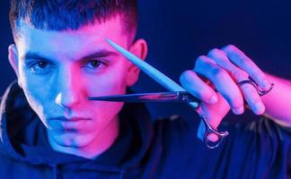 Holds scissors. Young barber with work equipment standing in the studio with neon lighting photo