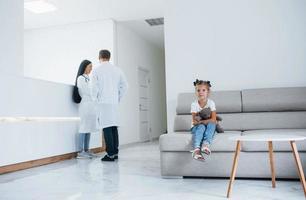 Doctors behind. Cute little girl with teddy bear in hands sits in waiting room of hospital photo