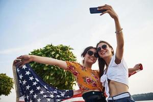 árbol verde en el fondo. dos mujeres alegres patrióticas con la bandera de estados unidos en las manos haciendo selfie al aire libre en el parque foto