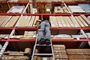 Storage worker stands on the ladder in uniform and notepad in hands and checks production photo