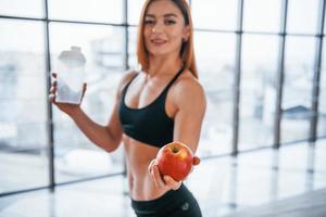 Sportive woman with nice body stands indoors with bottle of water and fresh apple in hands photo