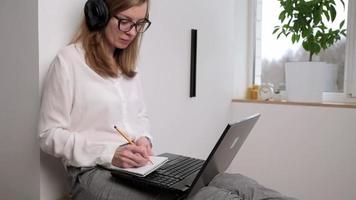 Remote work. Woman in headphones makes notes in notebook and print at laptop keyboard. Distance education and e learning concept video